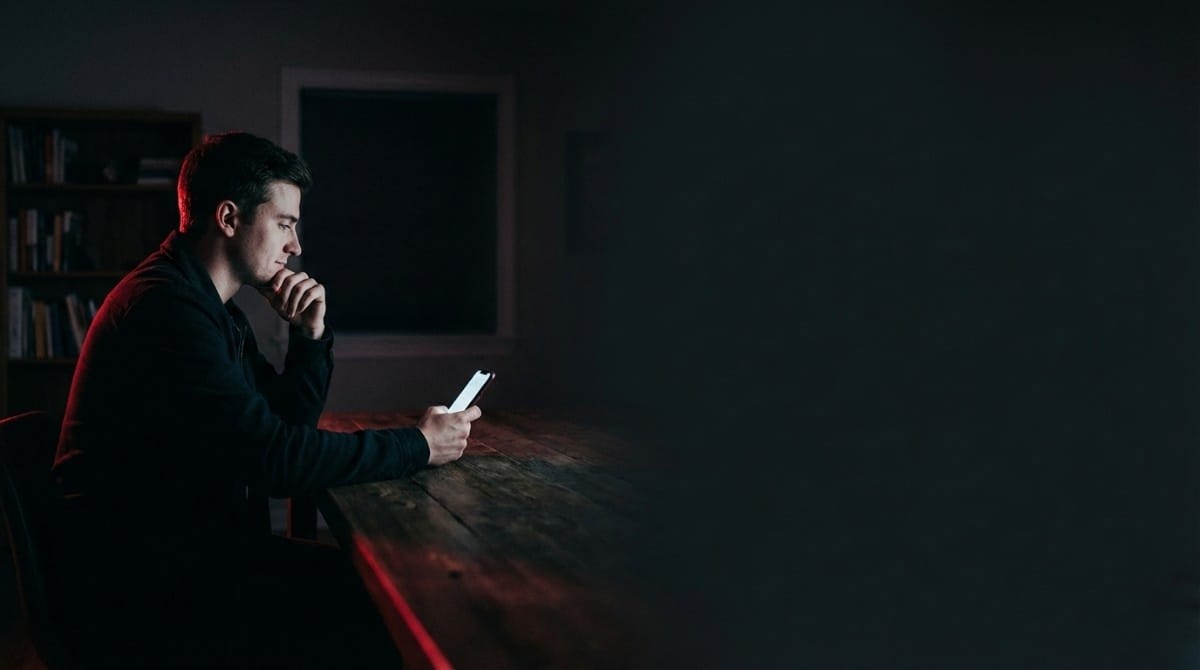 Man sitting alone at a table at night, looking at his phone thoughtfully with a serious expression