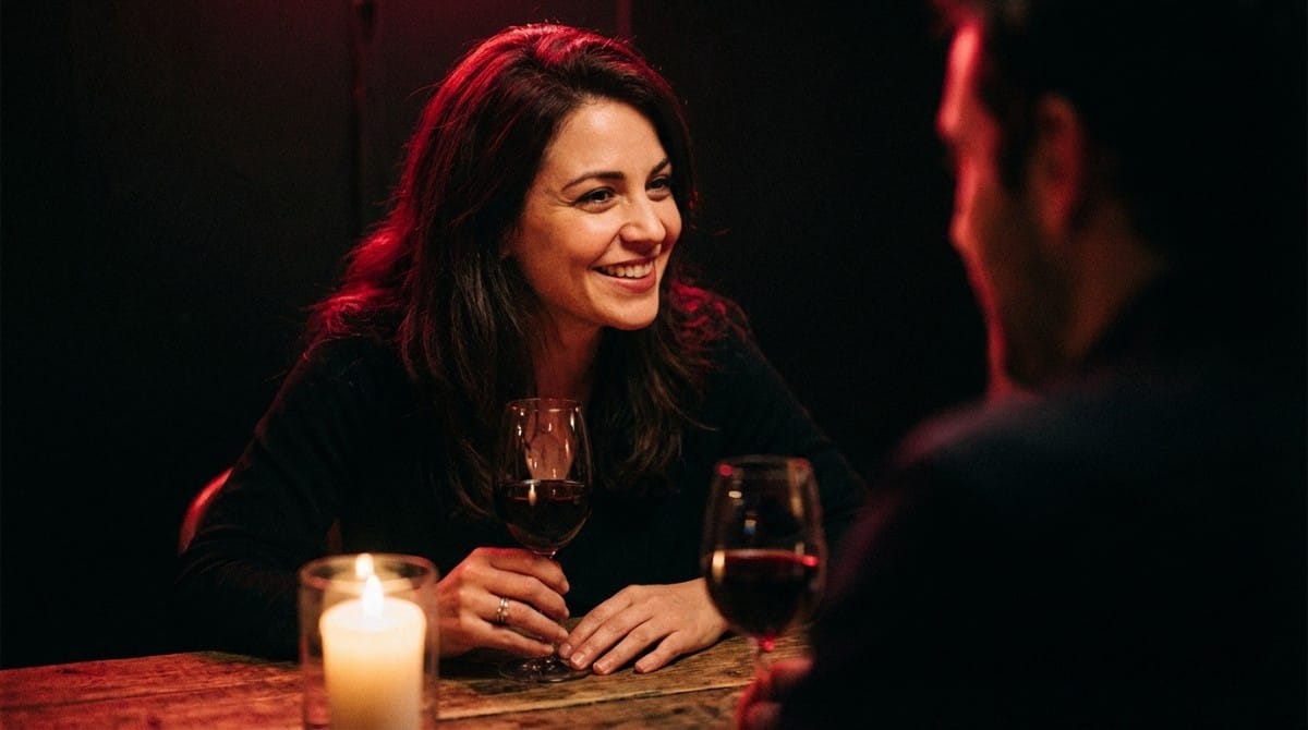 Woman smiling warmly during a candlelit dinner date, holding a glass of wine in a dimly lit romantic setting.