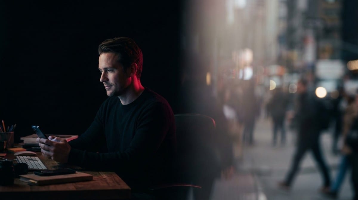 Man sitting at a desk using a smartphone in a dimly lit workspace with city lights visible through a window at night
