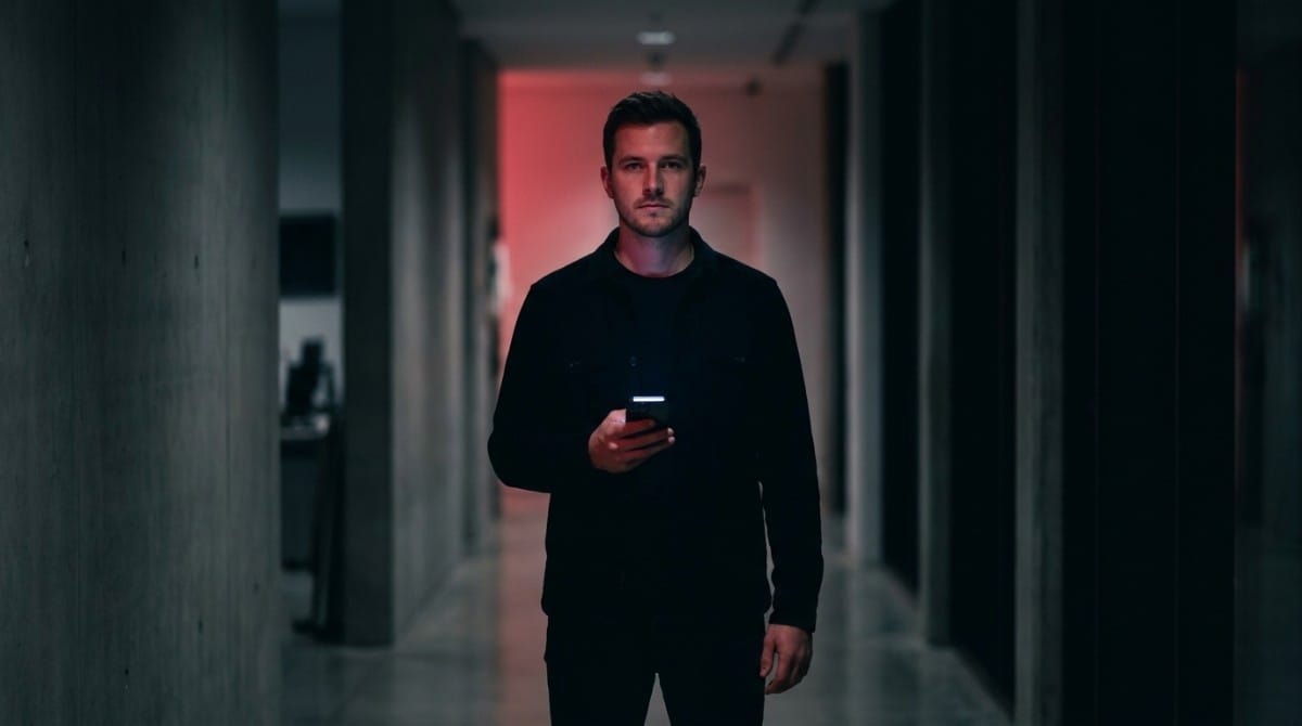 Man walking in a dark corridor holding a smartphone, illuminated by soft red and ambient lighting.