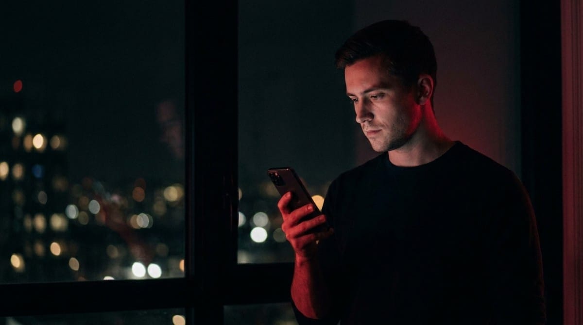 Man standing by a window at night looking at his smartphone with city lights bokeh in the background and red side lighting
