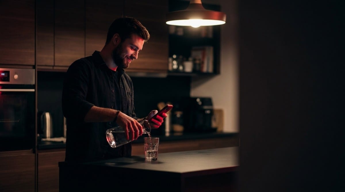 Man in a modern kitchen pouring water into a glass while looking at his smartphone with warm lighting and a cozy interior background.