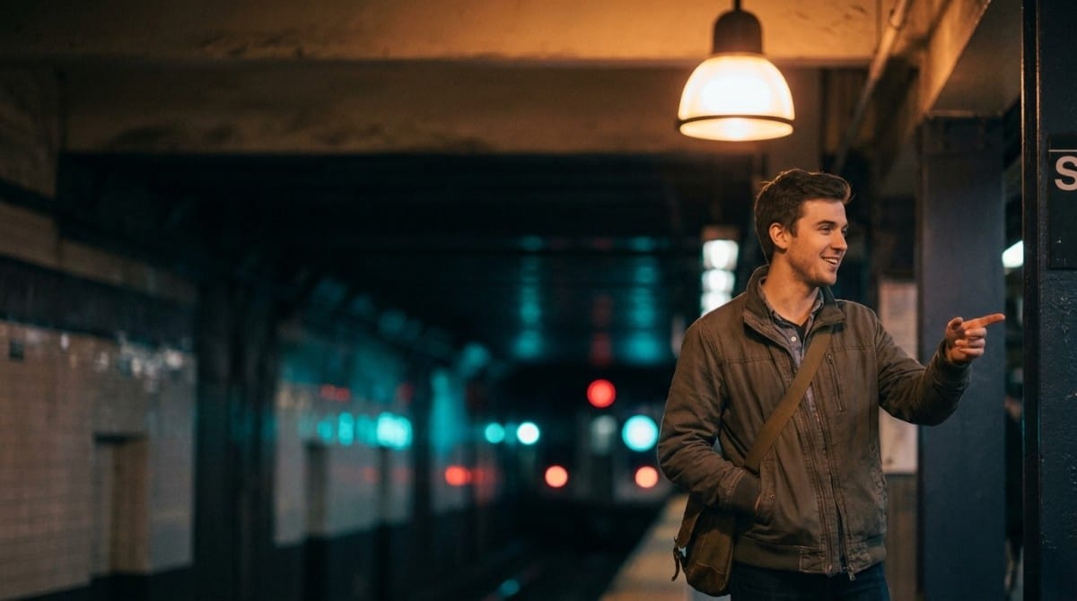 Man standing on a subway platform pointing ahead, with blurred train lights in the background.