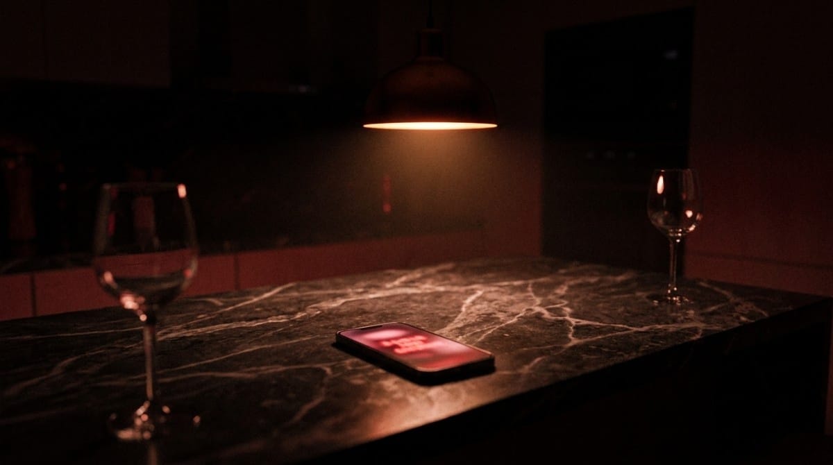Dimly lit kitchen counter with a smartphone displaying a red notification glow between two wine glasses under warm pendant lighting.