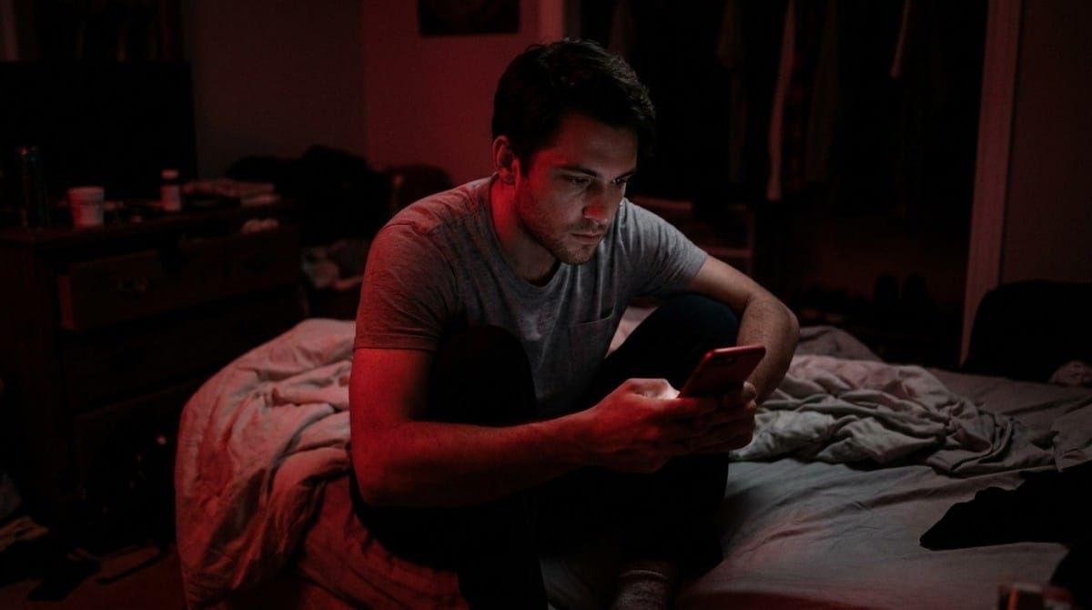 Young man sitting on a bed at night while looking at his smartphone in a dark room with red lighting.