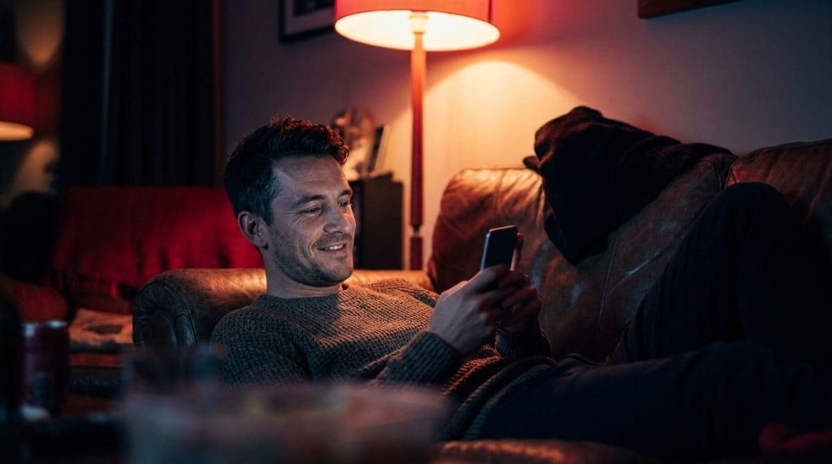 Man relaxing on a couch at night while smiling and looking at his smartphone in a dimly lit living room.