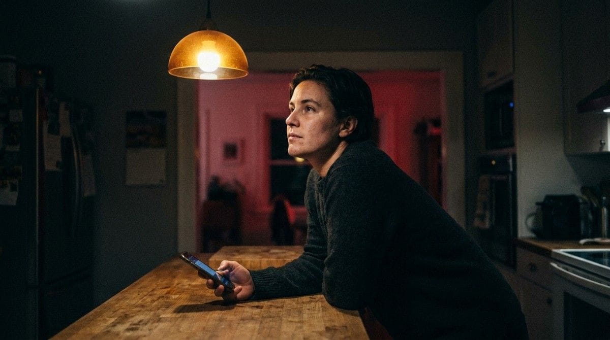 A person leans on a kitchen counter holding a smartphone, looking thoughtful and concerned in a softly lit home at night.
