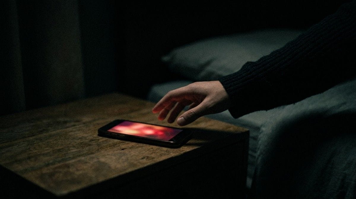A hand reaches toward a glowing smartphone on a bedside table in a dark room, representing late-night checking, anticipation, and emotional preoccupation
