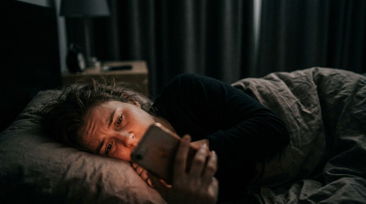 Person lies awake in bed at night staring at a smartphone with a worried expression.