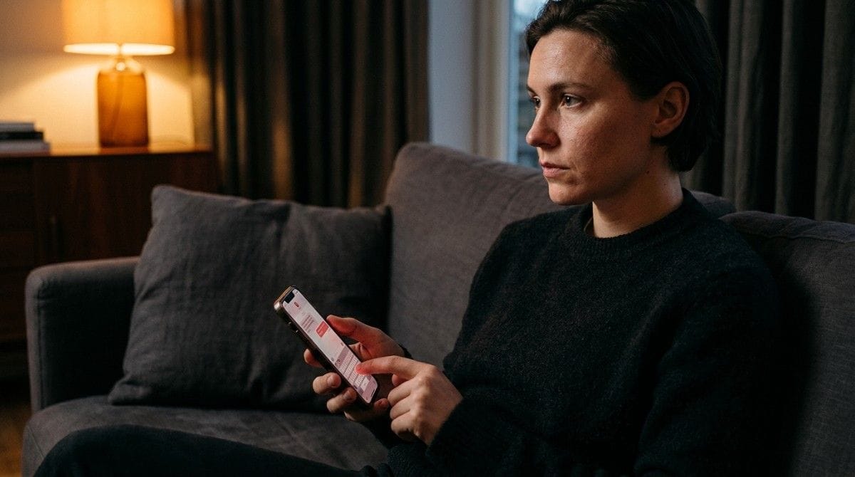 Person sitting on a couch looking at a smartphone with a serious expression in a cozy living room.