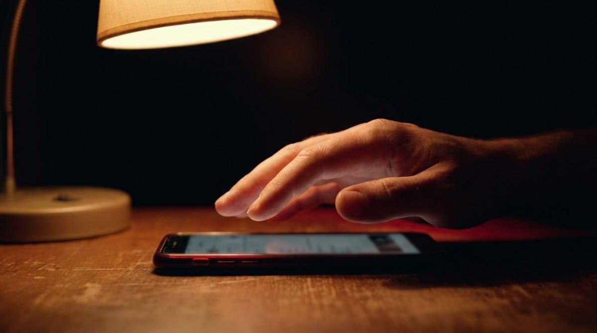 Close-up of a hand reaching toward a smartphone on a wooden table under a warm desk lamp.