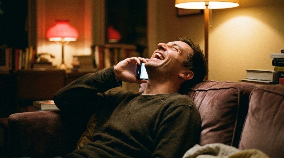 Man laughing while talking on the phone on a cozy living room sofa at night.