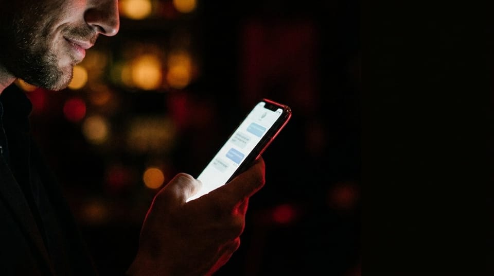 Close-up of a man smiling while reading chat messages on a smartphone in a dimly lit bar or lounge.